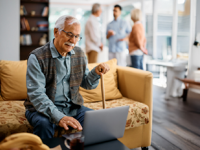 Usuario senior con un portátil en una clínica o residencia. En segundo plano y desenfocados un médico hablando con una pareja más joven