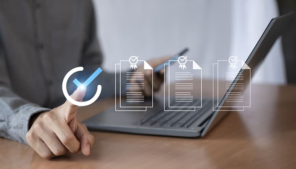 Una persona con traje utiliza un lápiz digital para marcar elementos en una lista de verificación virtual, con un portátil y una tableta sobre el escritorio.
