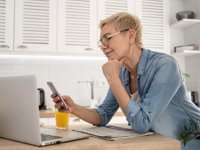 Usuaria en la mesa de la cocina con el portátil, el periódico y consultando el móvil