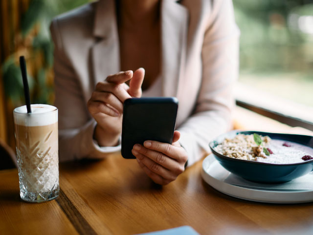 Detalle del busco de una usuaria en un restaurante, con un plato de comida y una bebida, haciendo una operación a través de su móvil