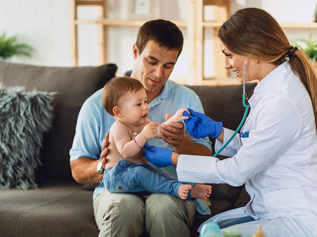 Una médica examina a un bebé que está en la piernas de su padre